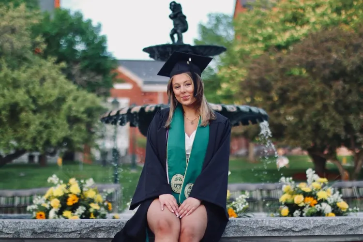 student wearing cap and gown sitting on a UVM campus fountain