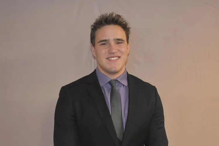 young man with short, spiky light brown hair, wearing dark gray suit and lavender shirt with tie, standing in front of wall