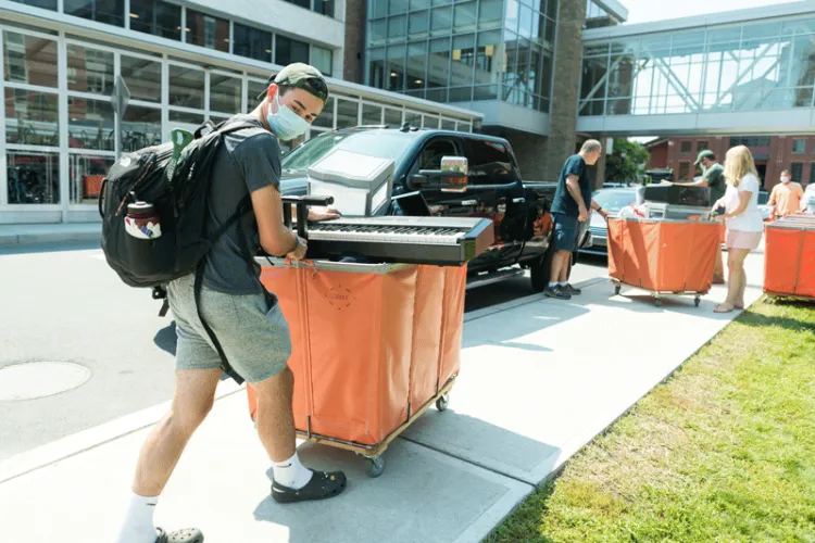 Student carefully wheels a cart with a keyboard balanced on top from car to residence hall
