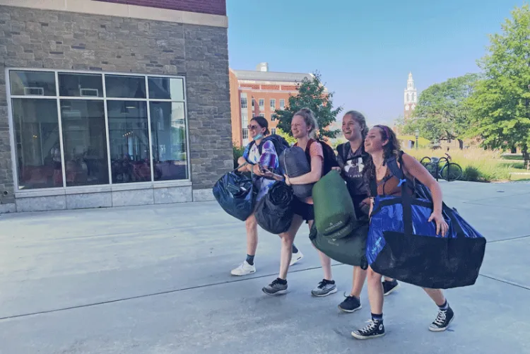 Four students carry large bags as they return from a camping trip.