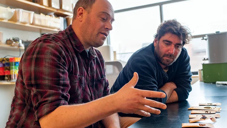 two men talking and gesturing with their hands in front of envelopes of seeds across a lab bench