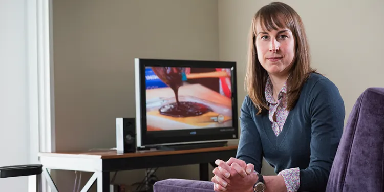 Lizzy pope sitting in a chair turned towards the camera with her hands clasped and a tv in the background