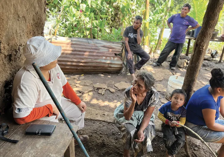 A group of people sitting outside on and around a dirt-like floor near a shelter all looking in different directions