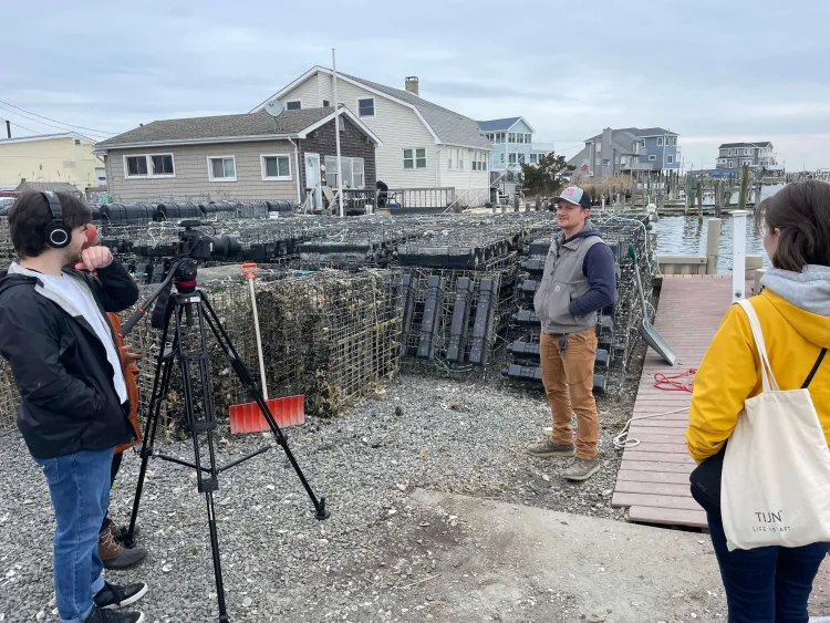 Person being filmed while talking next to stacked lobster traps on a dock. Two people listen; one holds a camera on a tripod. Houses are visible in the background.
