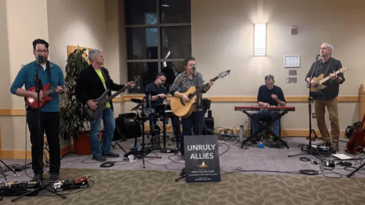 Six individuals in a band playing instruments behind a sign with the band name.