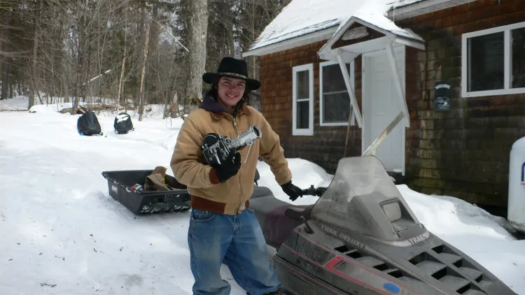 young man with brimmed hat and tan jacket holding photo equipment and standing by snowmobile in front of snowy yard and red house
