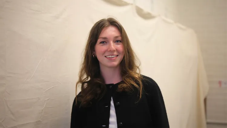 young woman with long light brown hair and black sweater over white shirt standing in front of canvas backdrop and smiling