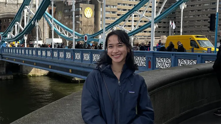 young Asian person with long dark hair wearing blue jacket standing in front of bridge in urban setting