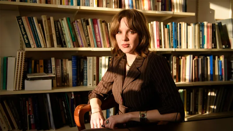 Young woman with brown hair wearing brown suit sitting at table in front of bookshelf
