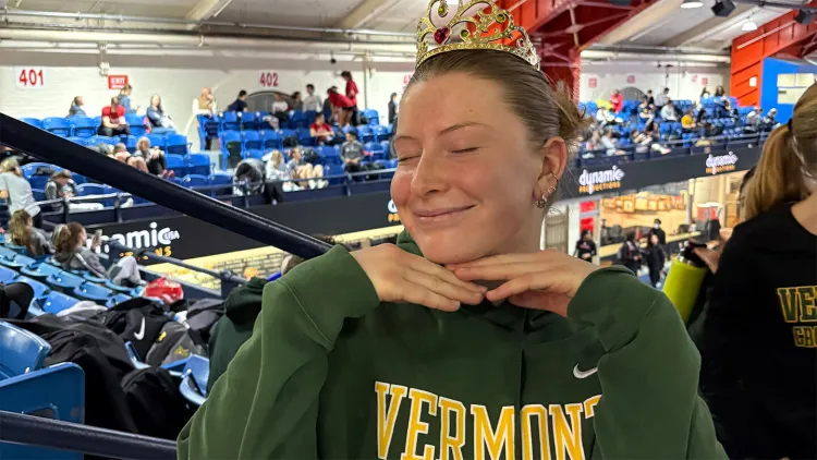 Young woman with brown hair pulled back in bun, wearing green UVM t-shirt with "Vermont" spelled out in yellow, standing in gym bleachers with eyes closed and hands under chin
