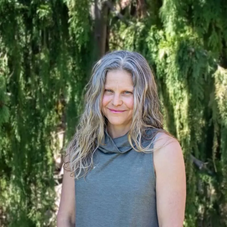 Rachelle Gould headshot, she is wearing a grey sleeveless shirt and is standing in front of a green, forested background