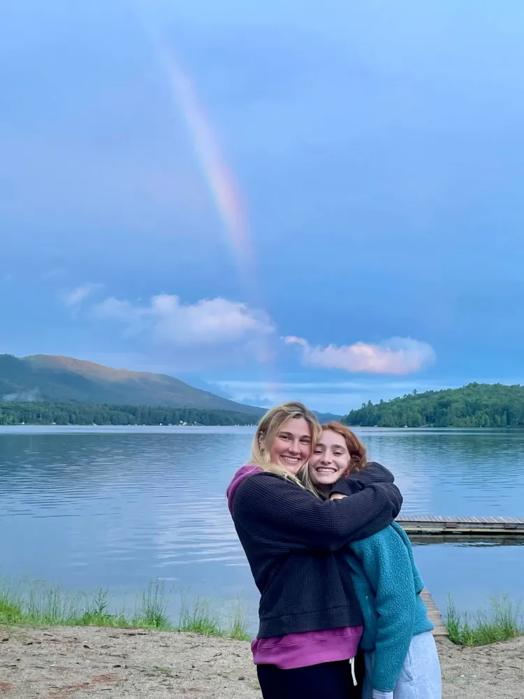 Chloe and a friend in front of a lake with a rainbow in the sky.