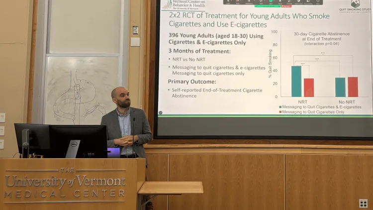  man presnting at a podium with a big screen showing his research in the background