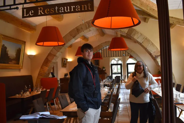 A person in a black jacket and a woman in a white sweater stand inside a restaurant with stone arched ceilings and a sign that reads "Le Restaurant."