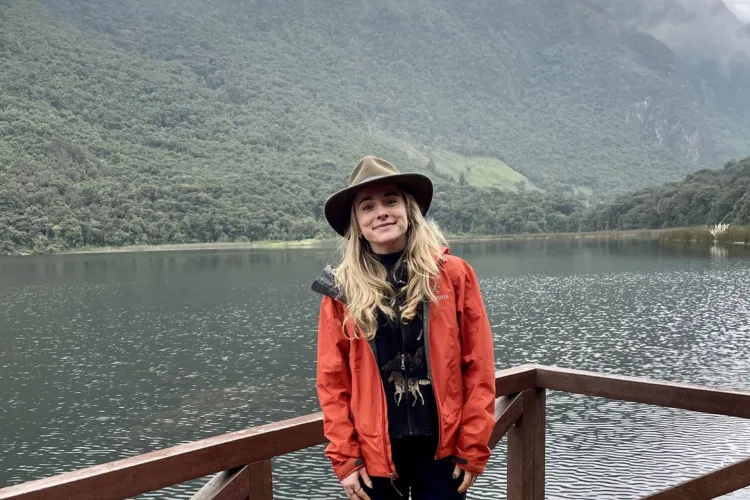 A smiling woman wearing a hat and a red jacket stands on a wooden deck in front of a calm lake and a green, tree-covered mountain under a cloudy sky.