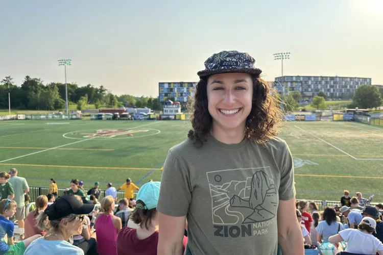 A person wearing a patterned hat and a t-shirt with a Zion National Park logo smiles in a stadium. The background shows a crowd in the stands and a sports field.