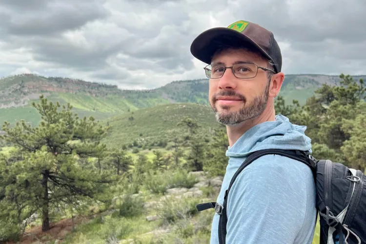 A man with glasses and a beard, wearing a baseball cap and backpack, stands outdoors with green hills and trees in the background under a cloudy sky.