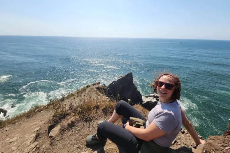A smiling woman sits on a rocky cliff overlooking a blue ocean.