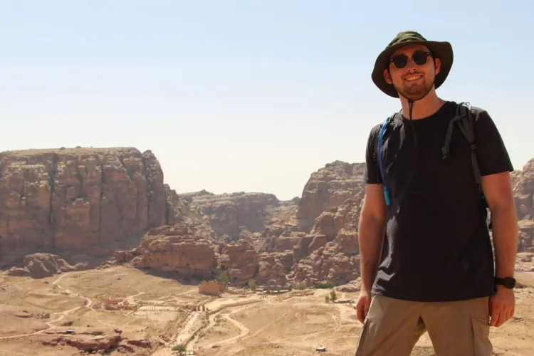 A person wearing a sun hat and sunglasses, with a backpack, stands on a rocky overlook, smiling at the camera. In the background is a vast, arid canyon landscape with a path winding through it.