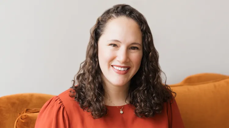 Smiling woman with curly brown hair wearing an orange top and a gold necklace.