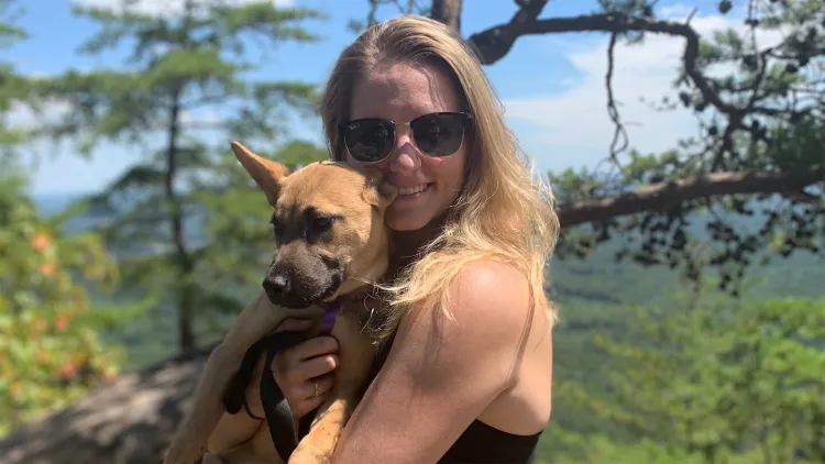 A person holds a puppy while standing on a mountain with a forested background.