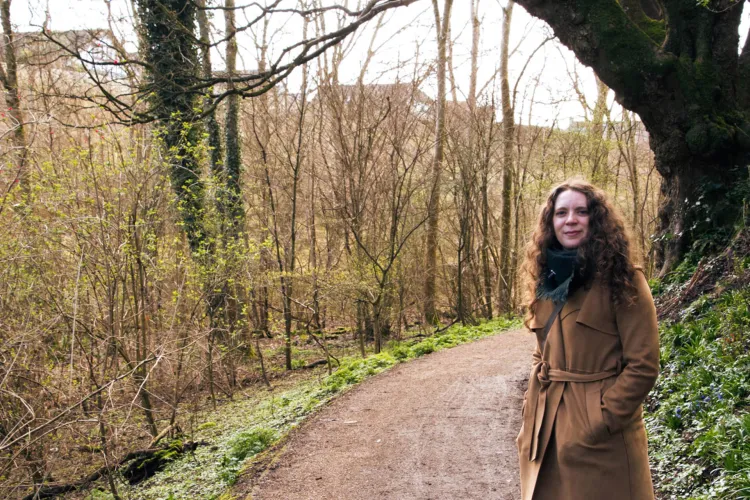 A woman wearing a tan trench coat stands on a dirt path in a wooded area. She is looking at the camera with a slight smile.