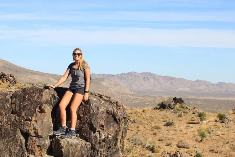 A smiling woman in sunglasses, shorts, and a t-shirt is sitting on a large rock in a desert landscape. In the background are hills and sparse vegetation under a clear blue sky.