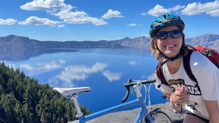 A cyclist smiles with her bike in front of a stunning blue lake with mountains in the background.