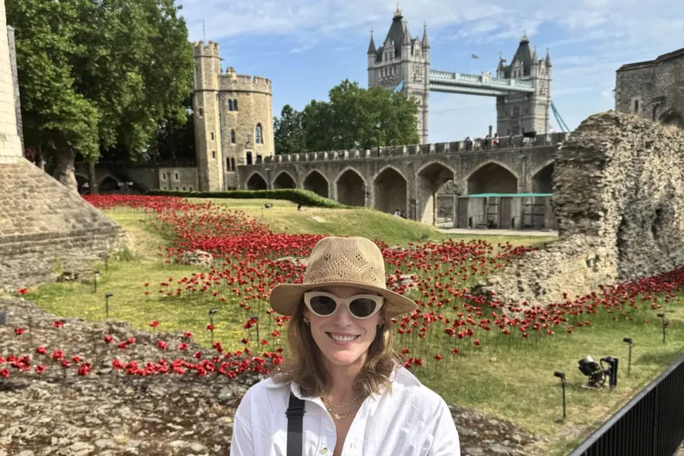 A person is smiling at the camera, wearing a straw hat, white sunglasses, and a white shirt. Behind them is a large field of red poppies. In the background, there is a stone fortress and the Tower Bridge.