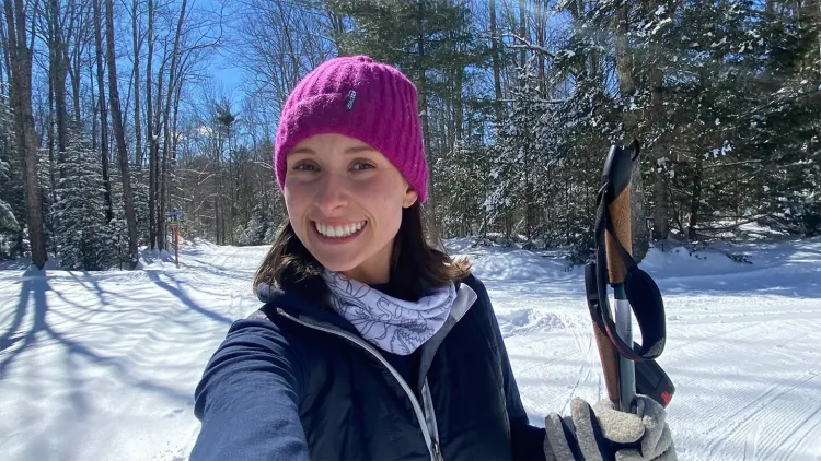 Smiling person taking a selfie outdoors in a snowy forest, wearing a bright pink knit hat, scarf, and winter jacket, holding a ski pole