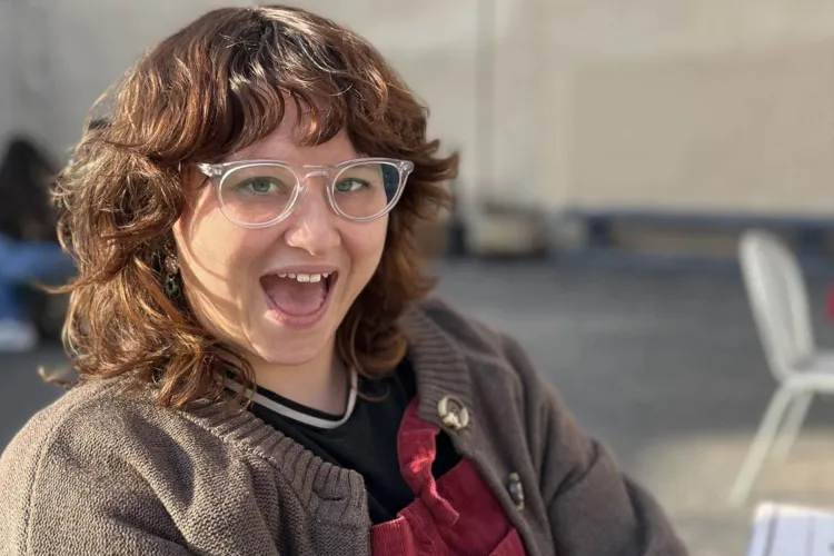 A person with glasses smiles with an open mouth, wearing a cardigan over a dark shirt and a red apron. The background is a blurry outdoor setting.