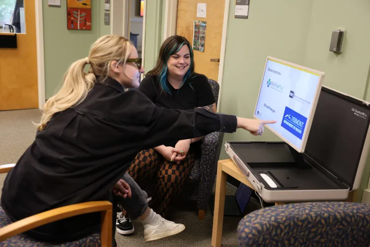 Two women sitting in front of a small pop-up monitor displaying logos for different digital health resources.