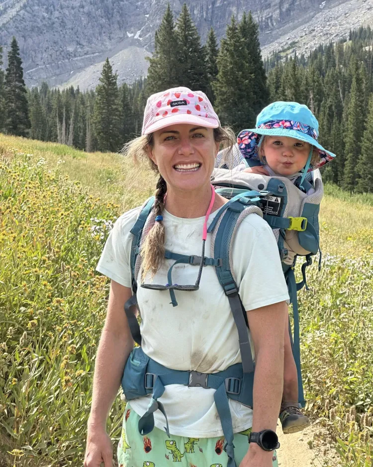Hilary Flint pauses on a hike. She is wearing a pink hat and a white shirt, and a baby-carrying backpack. A baby in a blue hat is in the backpack.