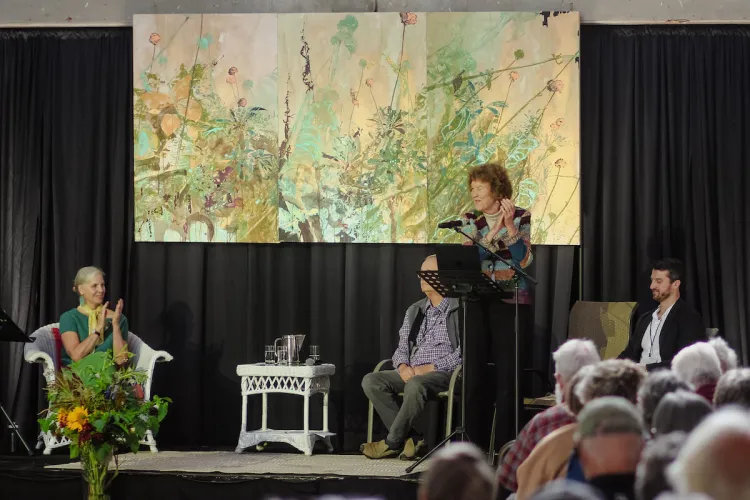 Four people on a stage, one is standing at a podium, the rest are sitting, behind the stage is a large painting