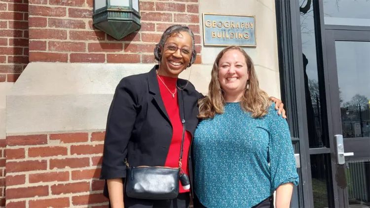 tall woman with glasses, black jacket, and red shirt standing next to woman with long blonde hair and blue sweater in front of brick building; both are smiling