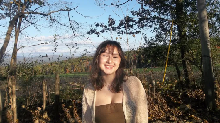 Young woman with long brown hair, brown top, and light-colored cardigan smiling at camera with autumn field and trees in background