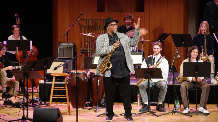 Jazz musician performing on stage with a saxophone alongside a student ensemble in a concert hall.