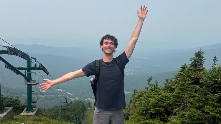 Young man with short, dark hair and navy blue t-shirt standing on misty mountain top with arms outstretched