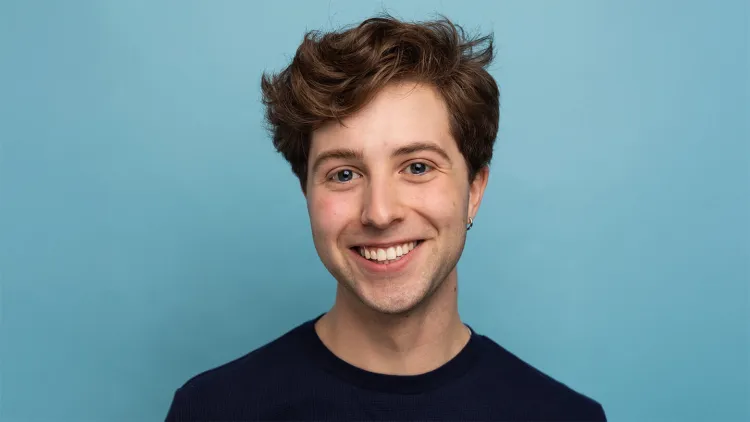 Young man with short, dark, curly hair and dark shirt standing in front of blue wall, smiling