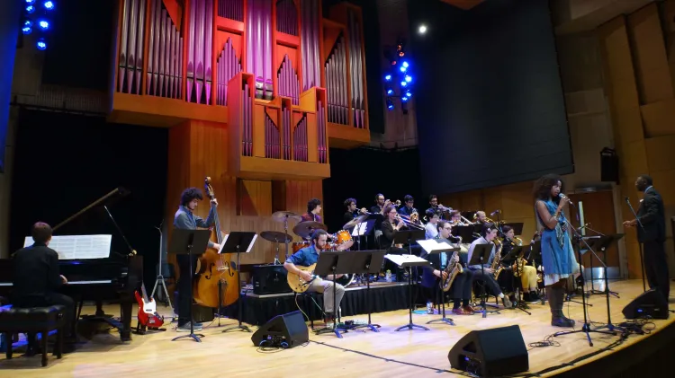 A jazz ensemble performs on a stage, featuring a singer in a blue dress, a drummer, a guitarist, a bass player, a pianist, and a large brass and woodwind section