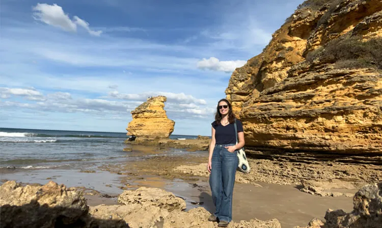 A graduate student posing for a photo on a beach with rocky cliffs.