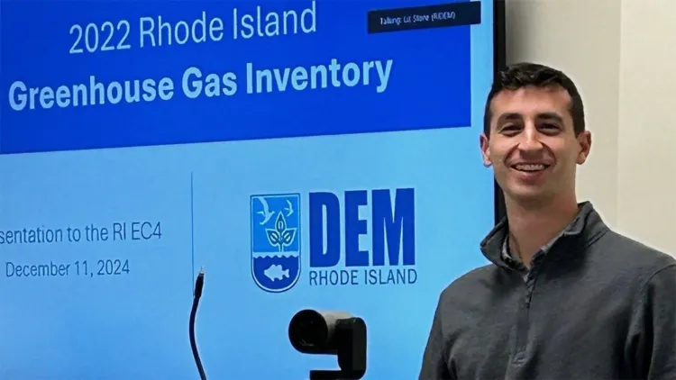man with short dark hair and grey pullover standing next to large projected slide that says "2022 Rhode Island Greenhouse Gas Inventory"