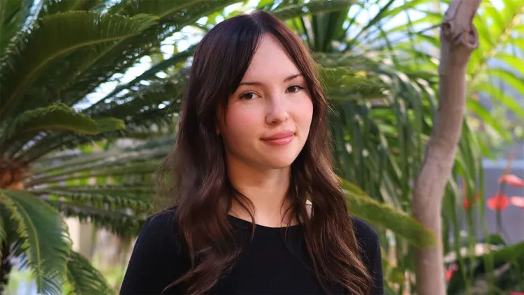 Young woman with long brown hair and black shirt standing in front of palm trees