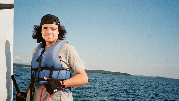 Person wearing a blue life jacket and cap standing on a boat, smiling with a lake and distant shoreline in the background.