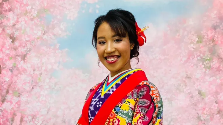 Young woman with dark hair wearing colorful traditional Japanese garment, smiling at camera while standing in front of blossoming cherry tree background