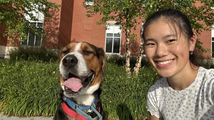 Young woman with dark hair and light top smiling at camera while crouched next to brown and white dog in front of brick building