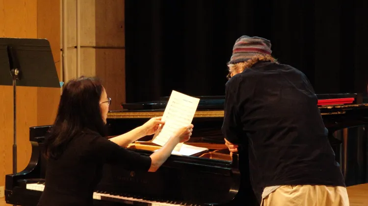 Person seated at a grand piano holding sheet music while talking with another person leaning beside the piano.
