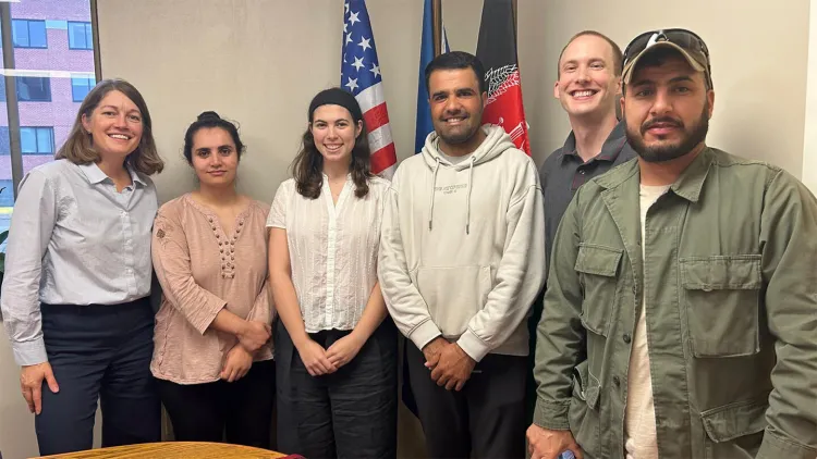 Six people standing in line in front of the U.S. and other flags; three women are on the left and three men are on the right
