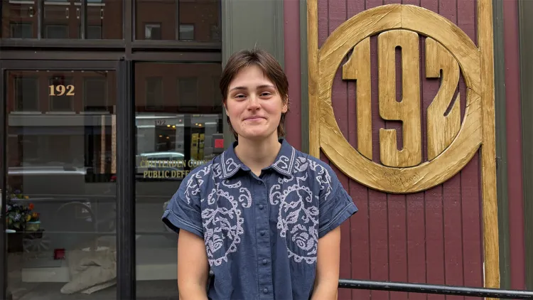 Young woman with short brown hair and blue shirt stands in front of a building with "Chittenden County Public Defender" on the door and "192" on the wall beside it