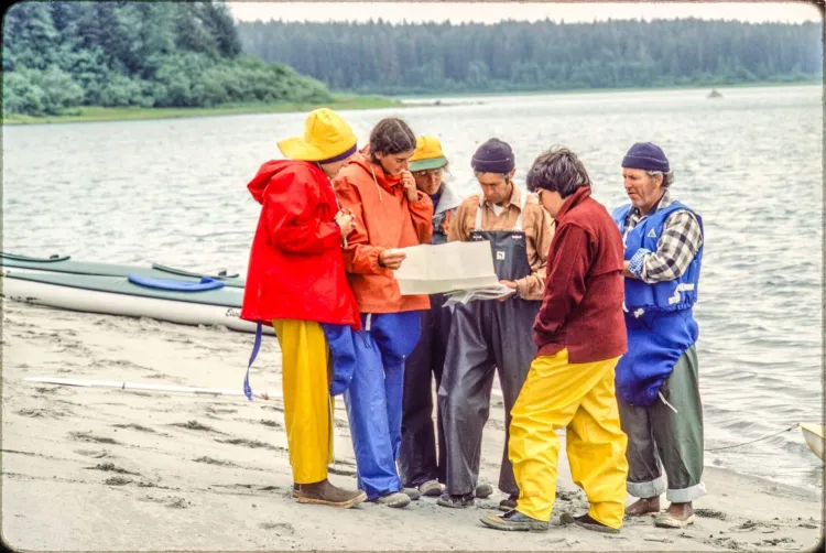 six people in colorful rain gear looking at a map together on a sand beach on a bay